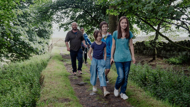 A family group of three children and two adults are walking along Hadrian's Wall. There are trees in full green leaf either side of them forming a canopy above.